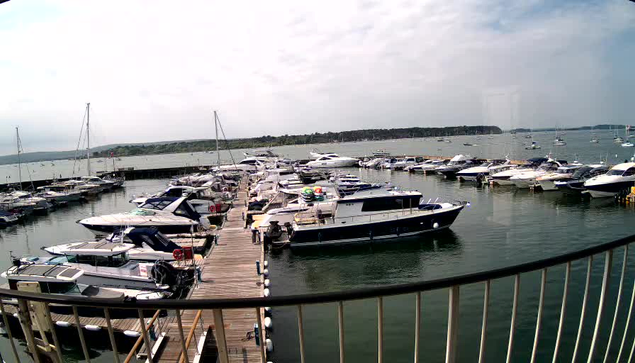 A view of a marina with numerous boats docked along a wooden pier. The water is calm, and there are additional boats visible in the distance on the open bay. The sky is partly cloudy, and a distant shoreline is visible on the horizon.