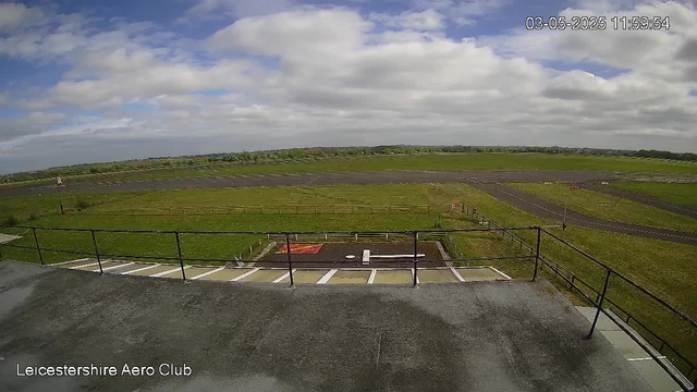 A wide view of an open grassy airfield under a partly cloudy sky. In the foreground, there is a concrete balcony with a black metal railing. Below the balcony, there are several parking spaces, some marked with white lines. The airfield has paved runways and taxiways visible extending into the distance, with a few small structures and signs. The top right corner displays a timestamp that reads "03-05-2025 11:59:54." The bottom left corner shows the text "Leicestershire Aero Club."