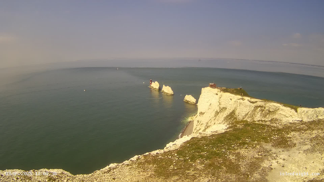 A panoramic view of white chalk cliffs overlooking the sea on a clear day, with a lighthouse and a small building on top of the cliffs. The water below is calm with a few boats visible in the distance, and the sky is mostly clear with some wispy clouds.