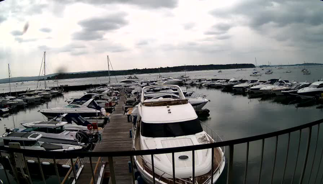 A marina with several boats docked along wooden piers, viewed from behind a black metal railing. The sky is cloudy and overcast, with multiple boats and yachts of various sizes anchored in the water.