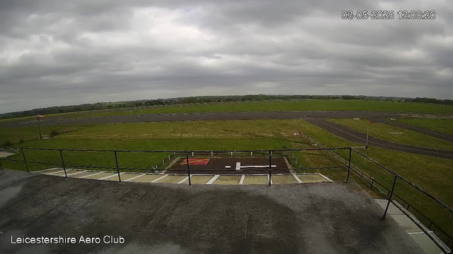 A view from a webcam overlooking an airfield with grassy fields and a runway stretching into the distance under a cloudy sky. The date and time are displayed at the top right corner, and "Leicestershire Aero Club" is written at the bottom left. There are parking or storage spaces with white lines and a railing in the foreground.