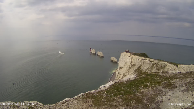 A view of white chalk cliffs along the coastline with a grassy top and a small building. The cliffs overlook the sea with several large rocks and sea stacks in the water below. The sky is cloudy, and a few boats are visible sailing near the cliffs.
