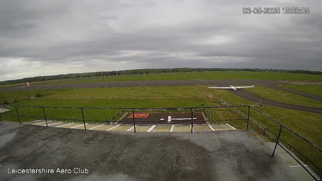A small white airplane is parked on a runway in an open field beneath a cloudy sky. In the foreground, there is a balcony or observation deck with a black railing. The image is tagged with "Leicestershire Aero Club" and the timestamp indicates the date and time as 03-05-2025 at 15:00:05.