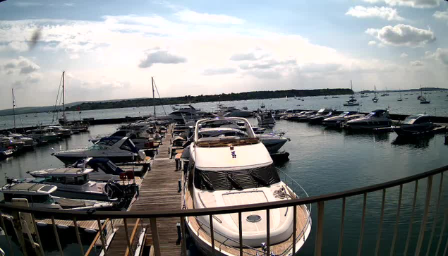 A marina filled with numerous docked boats and yachts. The boats are lined up along wooden piers extending into a calm body of water. The scene is under a partly cloudy sky with a distant horizon of land and additional boats visible in the background. The photo appears to be taken from a balcony or platform with a metal railing at the forefront.