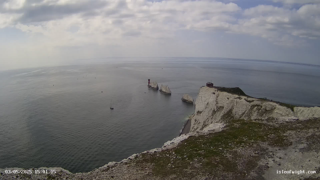 A panoramic view of white chalk cliffs rising from the sea, with a small structure and a lighthouse perched on top. Several boats are sailing near the cliffs, and a few people are walking along the edge. The sky is partly cloudy with patches of sunlight breaking through. The date and time are displayed in the bottom left corner, and a website URL is visible in the bottom right corner.
