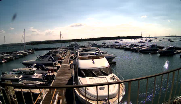 A marina with multiple boats and yachts docked at a wooden pier, under a partly cloudy sky with sunlight reflecting on the water.