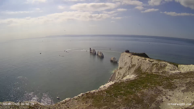 A panoramic view of a coastal area with white cliffs and a grassy top. There are large rock formations in the water near the cliffs, with boats and some seagulls visible in the sky and on the water. The sea is calm, reflecting the partly cloudy sky. A small structure is situated on the edge of the cliff, with a few people seen walking nearby. The photo is timestamped "03/05/2025 16:00:34" in the lower left corner.