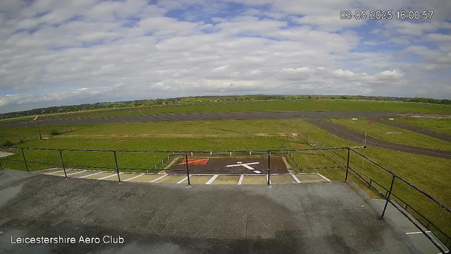 A wide view of an airfield with a grassy landscape and paved runways extending into the distance. The sky is partly cloudy with patches of blue. In the foreground, there is a concrete observation area with a black metal railing and marked parking spots. The bottom left corner displays the text "Leicestershire Aero Club," and the top right corner shows the date and time: "03-05-2025 16:00:57."