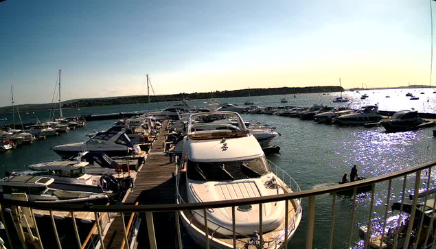 A marina filled with various boats and yachts docked along wooden piers, with calm water reflecting the sunlight. In the background, the horizon shows a distant shoreline with some trees and structures, and the sky appears clear with a soft gradient from light blue to pale yellow near the horizon.