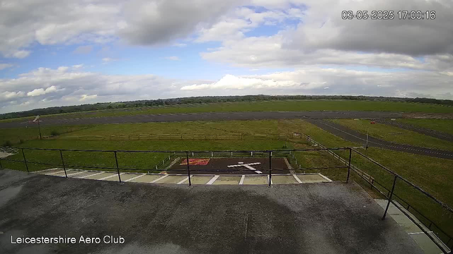 A view from a webcam at Leicestershire Aero Club shows an open grassy field with a small wind sock on the left side. The sky is partly cloudy with patches of blue visible. In the foreground, there is a concrete platform with parking spaces and a metal railing. The landscape extends into distant fields and tree lines under a mostly cloudy sky. The timestamp in the top right corner reads "03-05-2025 17:00:16".