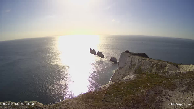 A view of white chalk cliffs descending into the ocean, with large sea stacks nearby. The sun is low on the horizon, reflecting brightly off the water, creating a shimmering effect. There is a small building or structure atop the cliff on the right side of the image. The sky is mostly clear with a few clouds. The scene is from a webcam, as indicated by the timestamp in the bottom left corner.