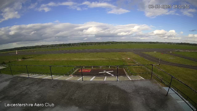 A wide view of an airfield with a grassy landscape stretching into the distance under a partly cloudy sky. The foreground shows a concrete area with a black railing, several parking spaces, and a marked helicopter landing pad with a white helicopter symbol. The timestamp in the top right corner reads "03-05-2025 18:00:50," and the bottom left corner displays the text "Leicestershire Aero Club."