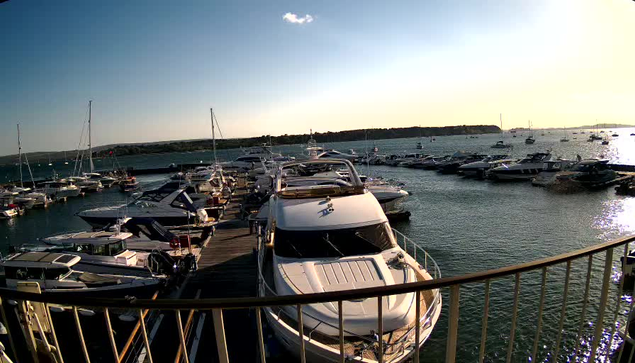 A marina filled with numerous boats and yachts docked at wooden piers. The water is calm, with a few ripples reflecting the sunlight. The sky is mostly clear with a few small clouds, and the horizon shows a distant landmass. A curved railing in the foreground runs along the edge of the dock area.