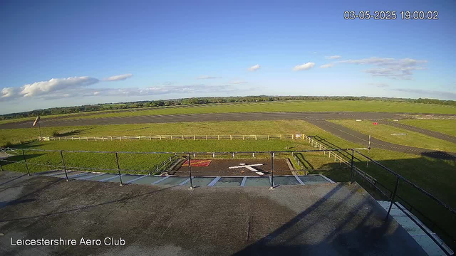 A wide view of an open grassy airfield with a runway stretching into the distance under a blue sky with scattered clouds. In the foreground, there is a rooftop area with metal railings and a small fenced section marked by an arrow. The image is labeled "Leicestershire Aero Club" in the lower left corner, and the date and time at the top right indicate it was taken on May 3, 2025, at 19:00:02.