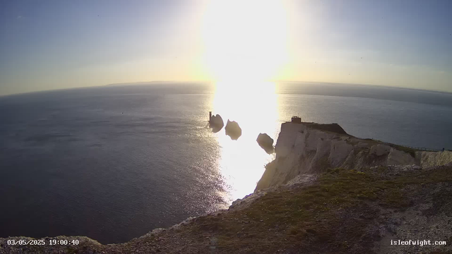 A view of a coastal cliffs with white chalk, overlooking the ocean during sunset. The bright sun is low on the horizon, casting a reflection on the water. There are a few large rocks near the cliff edge, and a small building or structure is visible on the right side of the cliff. The date and time displayed are "03/05/2025 19:00:40," and the website "isleofwight.com" is shown in the bottom right corner.