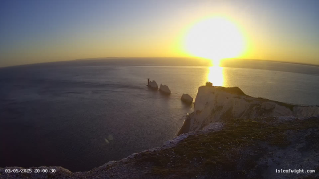 A sunset over the ocean near white chalk cliffs. The sun is low on the horizon, casting a bright reflection on the water. There are large rock formations and a small structure on top of the cliffs, with a path or railing along the edge. The sky is mostly clear with a gradient from blue to orange near the horizon. The timestamp shows "03/05/2025 20:00:30" and the website "isleofwight.com" is visible in the corner.