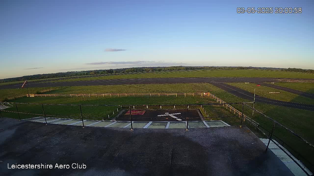 A wide view of an open grassy airfield with a small fenced area in the foreground. There are asphalt runways and taxiways stretching into the distance under a clear blue sky with a few scattered clouds. The image is timestamped "03-05-2025 20:00:58" in the top right corner, and "Leicestershire Aero Club" is written in the bottom left corner.