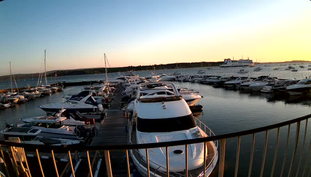 A marina filled with numerous boats and yachts docked along wooden piers, with calm water reflecting the vessels. The sky is clear with the sun setting, casting a warm glow over the scene, and a large boat is visible on the far right side of the water. A curved railing is in the foreground.