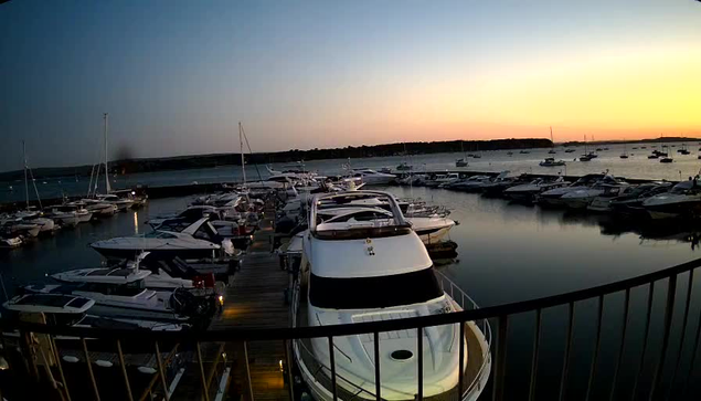A marina filled with various sailboats and motorboats docked along wooden piers during sunset. The sky is transitioning from light blue to warm orange hues near the horizon, reflecting softly on the calm water. A black railing is visible in the foreground, framing the scene.