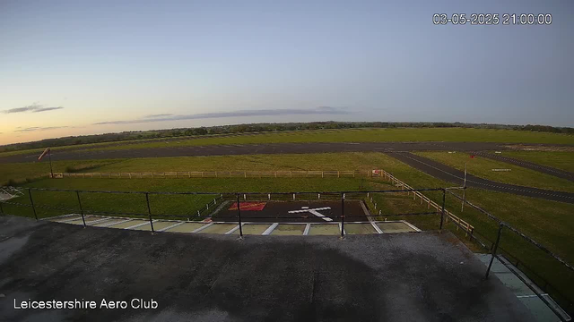 An empty runway and grassy field are visible under a clear sky during sunset. There is a small helipad marked with a red square and white helicopter symbol in the foreground, with a railing around it. A windsock is on a pole to the left, and a paved pathway runs alongside the grass. The image is labeled "Leicestershire Aero Club" in the bottom left corner, and the date and time "03-05-2025 21:00:00" are shown in the top right corner.