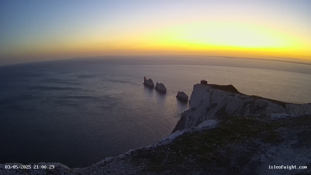A panoramic view of the ocean at sunset with white chalk cliffs in the foreground. Several rock formations and small structures are visible on the cliffs, and the sky is painted with soft hues of yellow and orange near the horizon. The scene appears calm and serene, with gentle ripples on the water's surface. The image is timestamped "03/05/2025 21:00:29" and is from isleofwight.com.