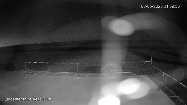 A black-and-white image taken by a webcam at night shows an empty, open outdoor area with a metal railing or fence running along the foreground. In the background, there is a vast open sky with no visible stars or clouds, and the horizon appears dark. The image has some out-of-focus bright spots or light reflections. The top right corner displays a timestamp: "03-05-2025 21:59:56," and the bottom left corner shows the text "Leicestershire Aero Club."