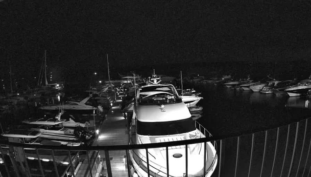 A black and white image taken from a webcam showing a marina at night. There are multiple boats and yachts docked, with some positioned along a floating dock. The boats vary in size and design, and the water is dark with reflections of the boats. A railing is visible in the foreground, indicating that the photo was taken from a viewpoint or balcony overlooking the marina.