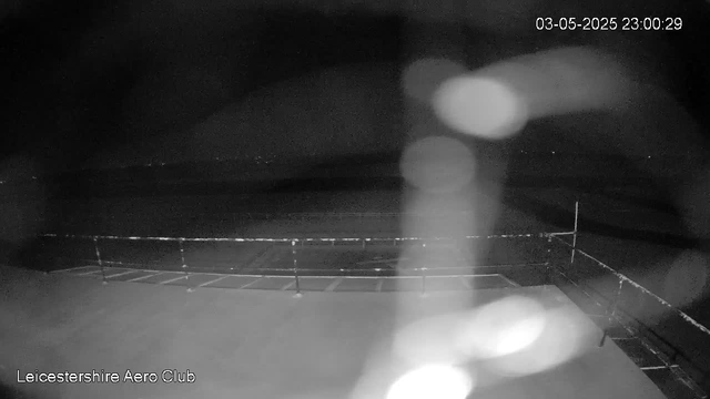A black and white image from a webcam showing an outdoor scene at night. There is a fenced area in the foreground, which appears to be part of an airfield or open space. The sky is dark, with a few small stars visible. The timestamp in the top right corner indicates the date and time as May 3, 2025, at 11:00:29 PM. The bottom left corner displays the text "Leicestershire Aero Club." There are some light reflections or lens flare effects in the image.