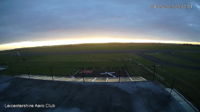 A wide view of an airfield during dawn or dusk with a cloudy sky. The image shows a runway, grassy areas, and a helicopter landing pad marked with a white helicopter symbol. In the bottom left corner, the text "Leicestershire Aero Club" is visible. The top right corner displays the date and time: 04-05-2025, 05:00:01. The perspective is from a camera mounted on a building or tower, looking out over the airfield.