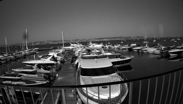 A black and white photo of a marina with numerous boats docked on either side. A wooden pier extends into the water, leading to larger yachts, with the horizon showing a distant shoreline under a clear sky.