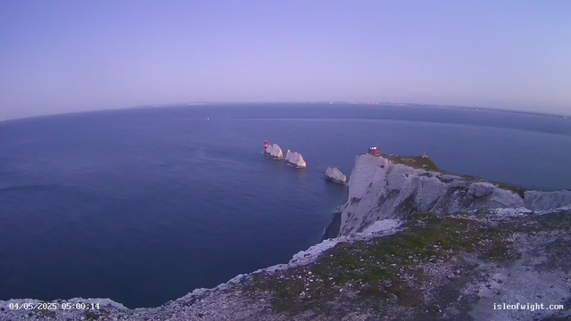 An aerial view of white chalk cliffs with green grassy tops extends into the distance over a calm, deep blue ocean under a clear, light purple sky. Several large white rocks are visible in the water near the base of the cliffs, with a small red and white lighthouse and a few structures on the cliffs. The photo appears to be taken early in the morning, as indicated by the timestamp "04/05/2025 05:00:14" in the lower left corner.