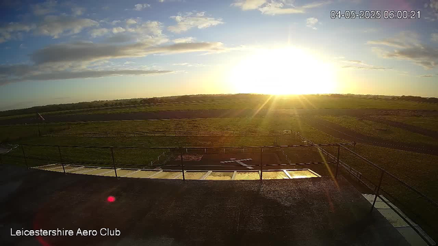 A view of a racetrack at sunrise, with the sun low on the horizon casting bright light and long shadows across the open field. The sky is partly cloudy with scattered clouds. In the foreground, there is a platform with a railing and what appears to be a paddock or preparation area. The setting is peaceful and illuminated by the early morning light. The text "Leicestershire Aero Club" is displayed in the bottom left corner, and the timestamp in the top right corner indicates the date and time as April 5, 2025, at 6:00:21 AM.