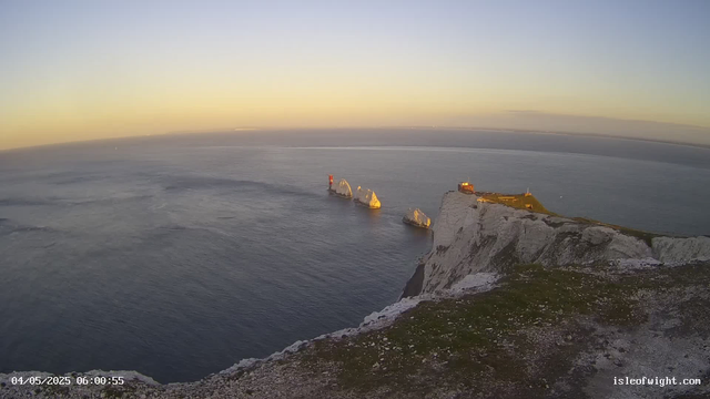 A sunset view of white chalk cliffs descending into the ocean, with several large sea stacks offshore. A red and white lighthouse stands on one of the rocks, and there is a small building on the edge of the cliff. The sky is clear with a warm glow from the setting sun. The timestamp reads 04/05/2025 06:00:55.