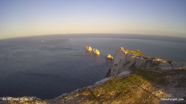 A panoramic view of a coastal landscape during sunrise, featuring tall white chalk cliffs with patches of green grass at the top. Several large rock formations are visible in the water near the base of the cliffs. A small building with a red top is perched on the cliff edge, and a lighthouse can be seen among the rocks. The calm ocean stretches out to the horizon with a few sailboats visible in the distance. The image has a timestamp of April 5, 2025, at 07:00:25.