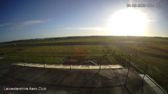 A wide view of an outdoor airfield shows a grassy area with a paved runway extending into the distance. The sky is mostly clear with a few clouds, and the sun is shining brightly near the horizon, creating glare. In the foreground, there is a platform with metal railings, and a small radar or antenna is visible. The timestamp in the top right corner indicates early morning on April 5, 2025. The bottom left corner has the text "Leicestershire Aero Club."
