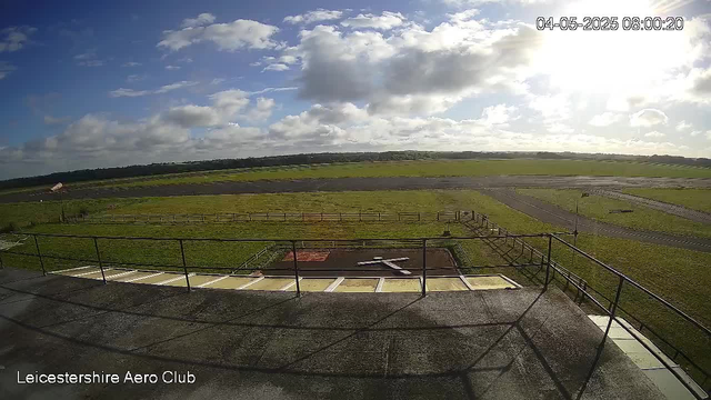 A view from a webcam shows an outdoor area with green grassy fields and a few paved taxiways or runways. The sky is partly cloudy with the sun shining brightly, creating a glare on the camera. A small satellite dish is visible on the left side, and a metal railing surrounds the edge of a platform or observation deck in the foreground. The bottom left corner has the text "Leicestershire Aero Club," and the top right corner indicates the date and time as "04-05-2025 08:00:20."