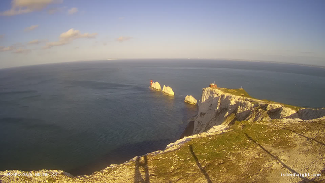 A panoramic view of white chalk cliffs overlooking the ocean, with a small building and a lighthouse on the edge of the cliff. Several large rocks are visible in the water near the base of the cliffs. The sky is mostly clear with a few scattered clouds.