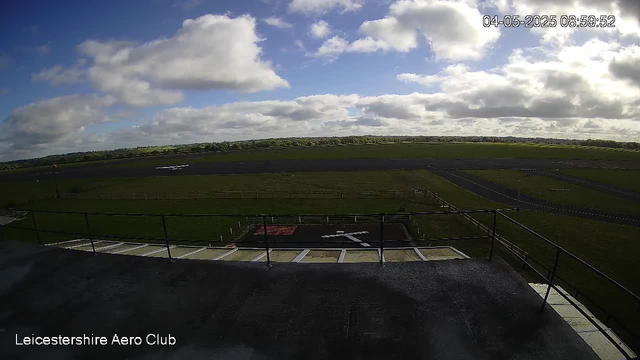 A view of an outdoor landscape under a partly cloudy sky, with green fields extending to the horizon. In the foreground, there is a balcony or rooftop area with a black railing and some small structures, possibly part of an airfield or aviation-related site. A taxiway or runway is visible in the middle distance, with a small airplane on the ground. The timestamp in the upper right corner shows "04-05-2025 08:59:52," and the bottom left corner displays the text "Leicestershire Aero Club."