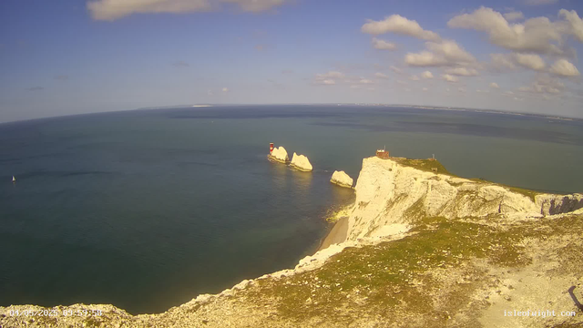 A wide view of the white chalk cliffs of Étretat in France, with a lighthouse on top of the cliff to the right. There are three prominent white rock formations in the sea near the cliffs, with one having a red and white lighthouse on top. The sky is partly cloudy with a few scattered clouds, and a sailboat is visible on the water to the left. The scene appears to be taken during daytime with clear weather.