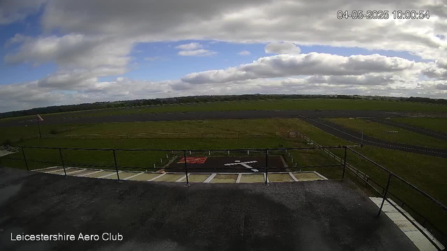 A view from a webcam showing an open grassy landscape with a runway or taxiway in the distance under a partly cloudy sky. The scene includes a rooftop or balcony with a black railing in the foreground, and some markings on the ground near the bottom of the image. The timestamp indicates the date and time as April 5, 2025, at 10:00:54. The text "Leicestershire Aero Club" is displayed at the bottom left corner.