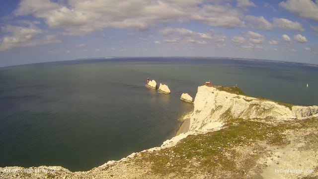 A view from a high vantage point of white chalk cliffs extending into the sea with white rock formations in the water. There is a lighthouse on the edge of the cliff near a wooden building, and several sailboats are visible on the water. The sky is partly cloudy with scattered white clouds.