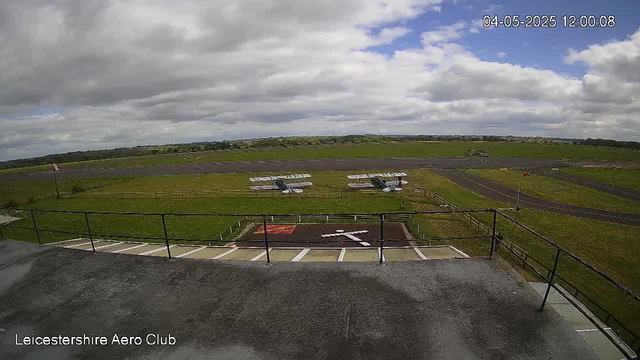 An outdoor view from a webcam at LeicesterShire Aero Club shows a cloudy sky with patches of blue. In the foreground, there is a concrete platform with a black railing, and behind it, a grassy area with small aircraft parked on the grass. The aircraft are white with blue and red accents. Further back, there are runways and taxiways, with some small buildings or structures visible in the distance. The time and date stamp in the top right corner reads "04-05-2025 12:00:08."