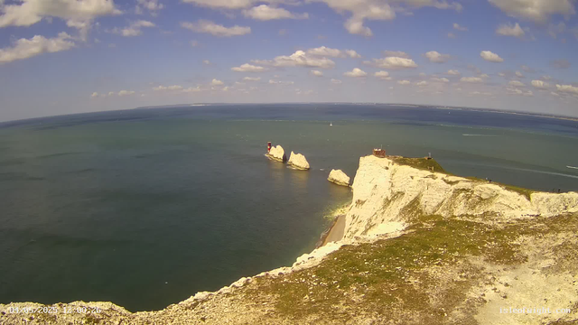 A panoramic view of a coastal landscape with white chalk cliffs extending into the sea. The cliffs are topped with green grass, and there are a few small structures on the highest points. In the water, large rock formations are visible near the base of the cliffs. The ocean is calm, with gentle waves, and several sailboats are seen in the distance under a partly cloudy sky.