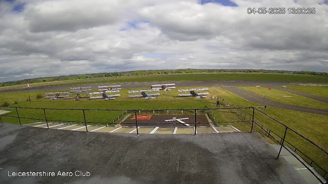 An outdoor scene at Leicester Aero Club shows a runway with several small aircraft parked on the grass, some with open doors or hatches. The sky is partly cloudy with patches of blue. In the foreground, there is a metal railing and a concrete platform with some parking spaces visible below. A person can be seen near the railing, and the field extends into a distant landscape with open green fields under a cloudy sky. The timestamp indicates the date and time as April 5, 2025, at 1:00 PM.