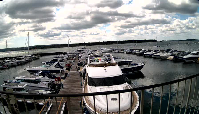 A view from a webcam showing a marina with multiple boats docked along a wooden pier. The boats vary in size and style, with some covered and others open. The sky is partly cloudy with patches of blue visible through the clouds. The water in the marina is calm, reflecting the boats and sky. In the background, there is a distant shoreline with trees. A curved railing is visible at the bottom of the image.