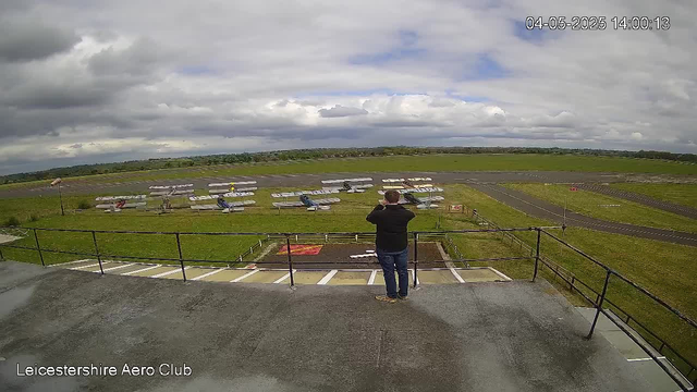 A person in dark clothing, wearing glasses, stands on an open viewing platform with a metal railing, looking out over a small airfield with several parked gliders or small planes. The airfield has runway markings and a grassy area surrounding the parked aircraft. The sky is mostly cloudy with patches of blue, and the scene is lit by natural daylight. The location is indicated as Leicesterhire Aero Club in the bottom left corner, with a timestamp showing the date and time as April 5, 2025, at 14:00.