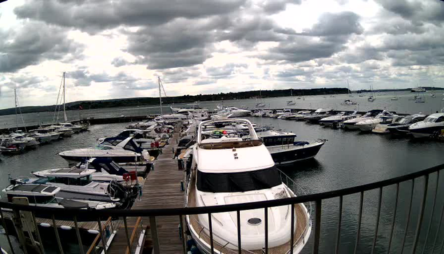 A view from a webcam showing a marina with multiple boats docked along wooden piers, under a cloudy sky.