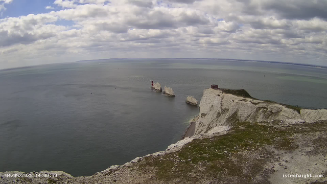 A view of white chalk cliffs extending into the sea, with several large rocks at the base, a structure on the cliff top, a red and white lighthouse on one of the rocks, and a cloudy sky overhead. The ocean surrounds the cliffs, and there's a grassy area on the cliff top.