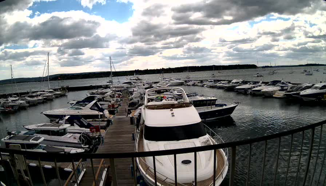 A marina with a variety of boats and yachts docked along wooden piers. The water is calm, and the sky is partly cloudy with patches of blue visible through the clouds. In the foreground, there is a white yacht seen from above, with the stern facing the viewer. A black railing curves along the bottom edge of the image.