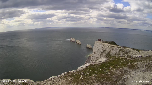 A wide view of the white chalk cliffs of Étretat in France, with a lighthouse on a rock formation in the sea and a cloudy sky overhead. The ocean stretches to the horizon, and a building is visible at the top of the cliffs.